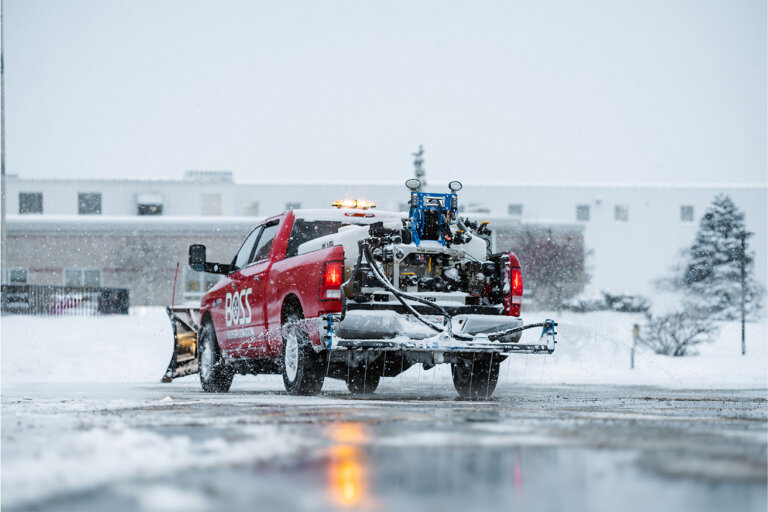 Red pick up truck in a snow covered parking lot with a snow plow on the front and liquid sprayer on the back.
