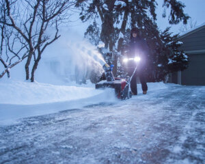 Woman using a Toro snow blower with headlights removing snow from a driveway at night