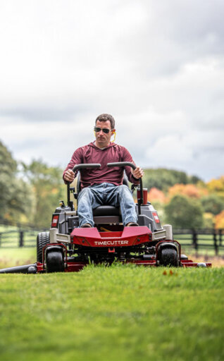 Man on a Toro TimeCutter zero turn riding mower cutting grass in a field with trees in the background