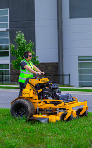 Woman in a neon green safety vest riding on a Wright stand on mower cutting grass