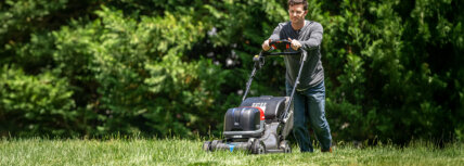 Man pushing a Honda lawn mower cutting high grass with green trees in the background