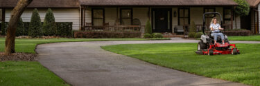 Woman on an red Exmark zero turn riding mower on a large lawn next to a winding driveway