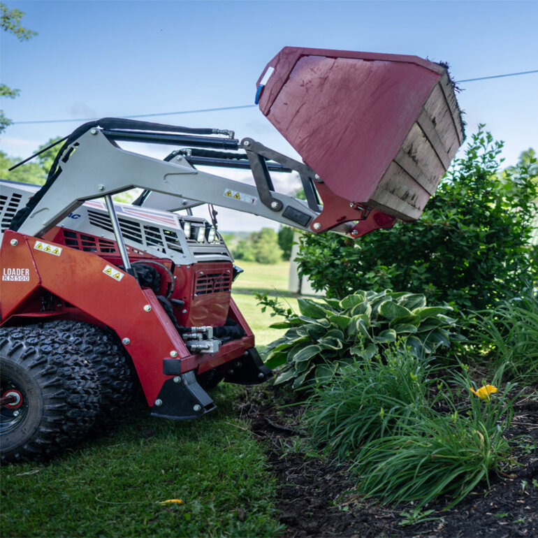 Vetnrac 4520 PRO tractor with power bucket attachment dumping mulch in a flower bed against a house